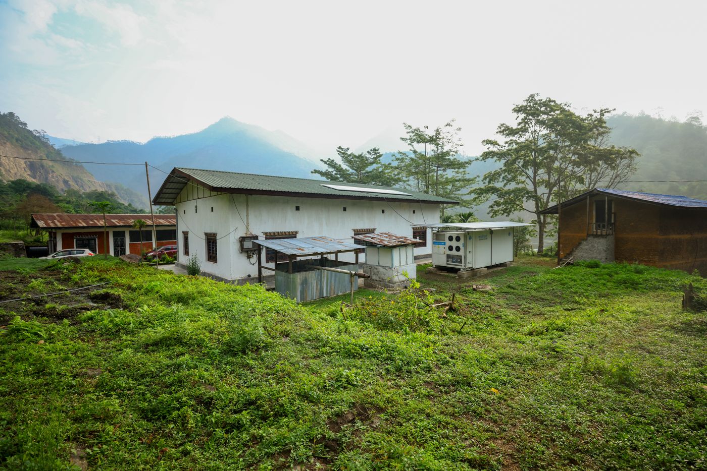 The solar cold storage unit beside a farm building with solar panels on its roof, below forested hills